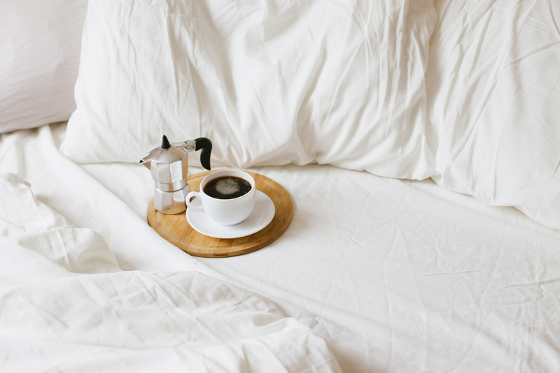 Cozy bedroom details: cup of hot fresh coffee and coffeemaker on wooden tray in bed with white linen.